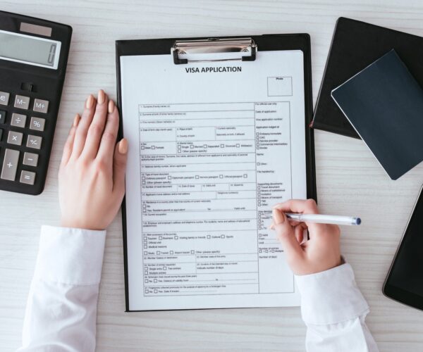 top-view-of-woman-holding-pen-near-document-with-visa-application-lettering-near-smartphone-with-e1692632864660.jpg